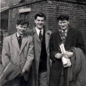Three young men in suits and overcoats standing in front of a building.