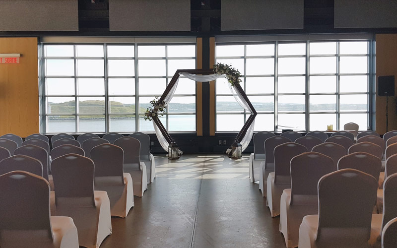 Wedding ceremony set with banquet chairs facing the floor to ceiling windows that overlook the harbour and island. A decorated hexagon arbor is set centre of the windows.
