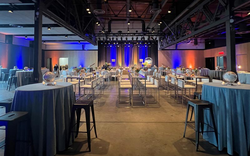 Tall clear bar tables and cruiser tables with blue linens have white bar stools for seating. The tables are decorated with blue linens, globe centerpiece and bud vases facing a stage with blue up lights.