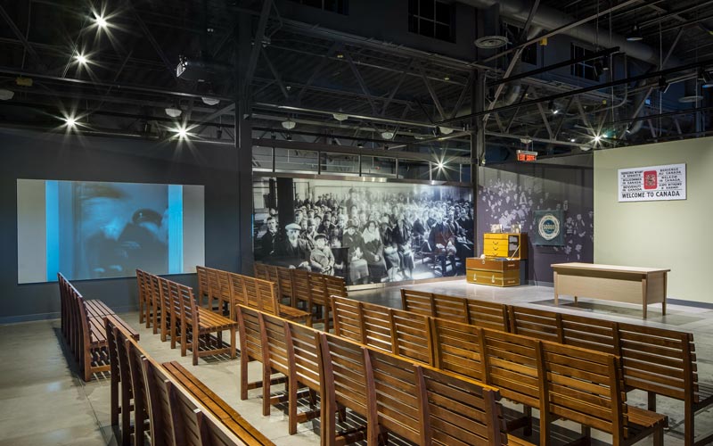 A Museum exhibition contains long wooden benches facing a table and a wall with a “Welcome to Canada” sign. To the left of the benches, there is a projection screen and a historic image of immigrants.