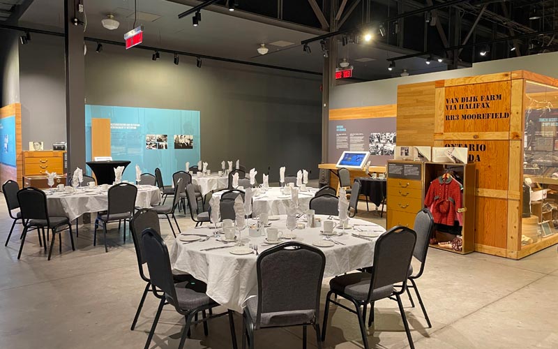 A banquet dinner set within a Museum exhibition space. Oval tables are set with banquet chairs, and the background includes a trunk and a suit case containing children’s clothing. 