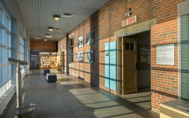A long narrow venue with floor to ceiling windows on the left overlooking the harbour. A brick wall has two doors, historic photos, and two benches for guests to sit. 