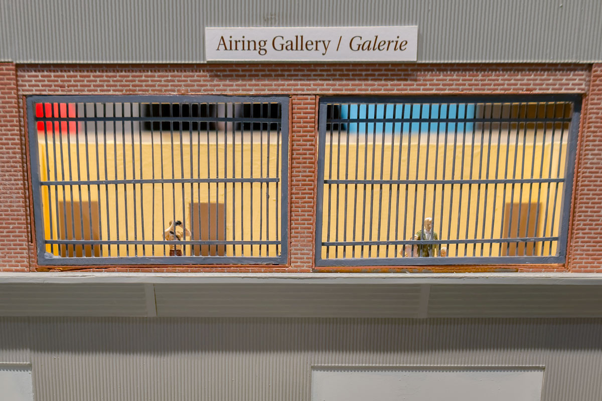 Model figurines of two light-skinned men looking out from behind metal bars on the second floor of a building.